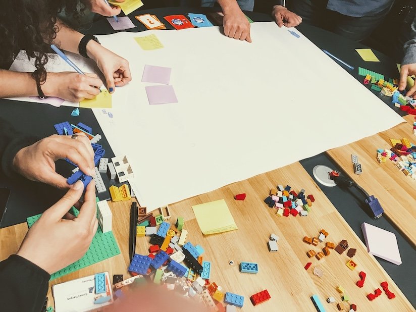 SysArt eğitiminden bir kare: Katılımcılar, büyük bir kağıt etrafında LEGO parçaları, kartlar ve post-it notlarıyla yaratıcı takım çalışması yapıyor. Görsel, oyun tabanlı öğrenme, iş simülasyonu ve çevik düşünme becerilerinin geliştirildiği interaktif bir eğitim ortamını yansıtır.