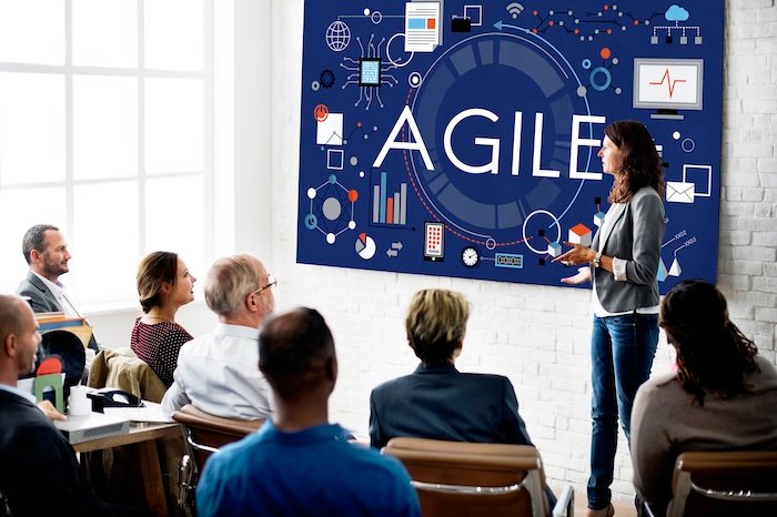Agile yazan bir tahta önünde toplantı yapan iş insanları görüntüleniyor.