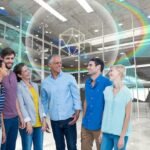 Smiling diverse group of people standing in a futuristic digital environment with holographic geometric shapes and rainbow light effects, symbolizing innovation and connectedness.
